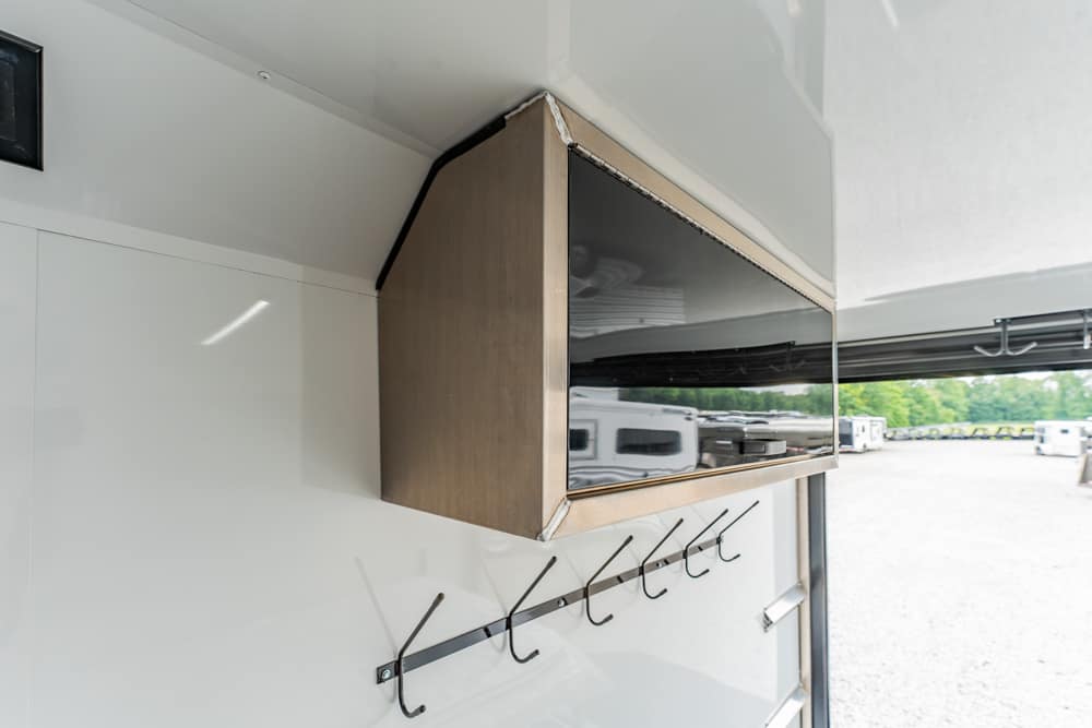 Flat-screen TV mounted in a wooden cabinet inside a white trailer, with coat hooks beneath it.