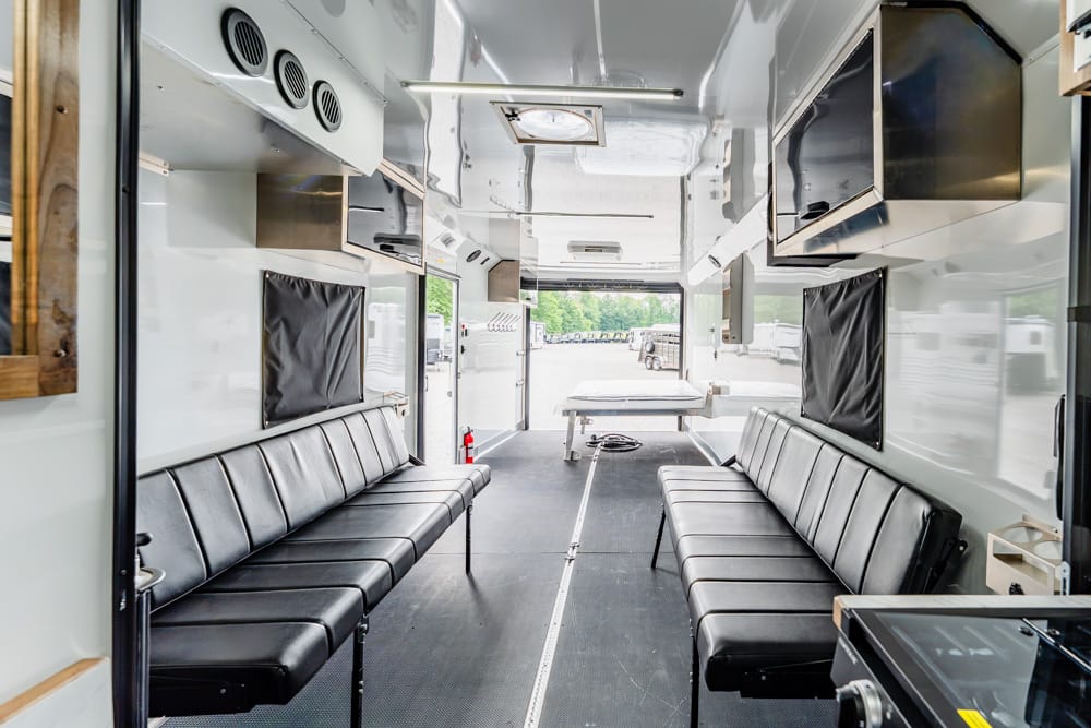 Interior of a mobile clinic/ambulance trailer with two long black bench seats along both sides and a central aisle at the rear table area in view.