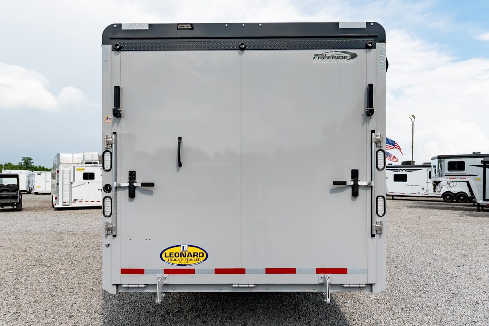 Rear view of a gray enclosed trailer with double doors, black latches, and a Leonard logo near the bottom; red/white reflective strips along the bumper on a gravel lot with other trailers in the background.
