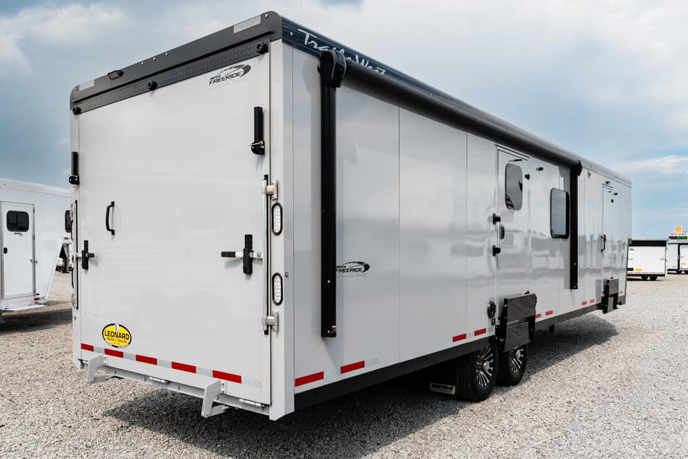 Rear and side view of a large white enclosed trailer with multiple entry doors and a rolled black awning along the top, parked on a gravel lot.