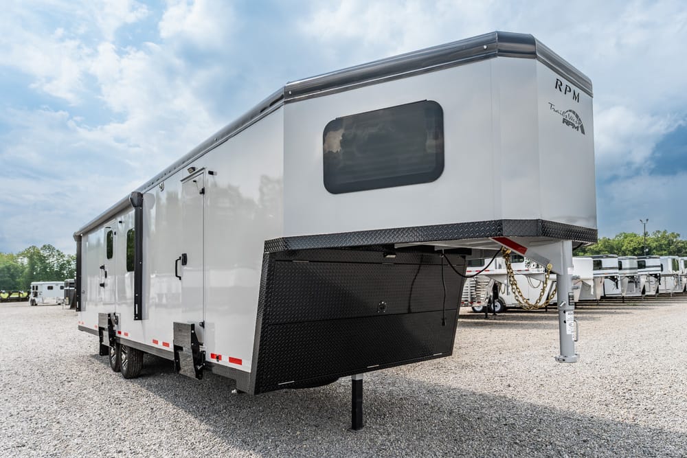 White 5th-wheel RV trailer with a raised gooseneck hitch and black diamond-plate storage box, parked on a gravel lot.