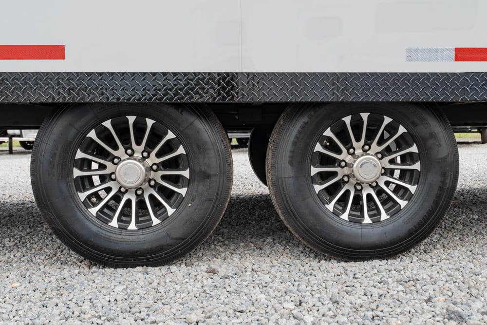 Twin trailer wheels with chrome rims under a white trailer on a gravel surface, side-by-side.