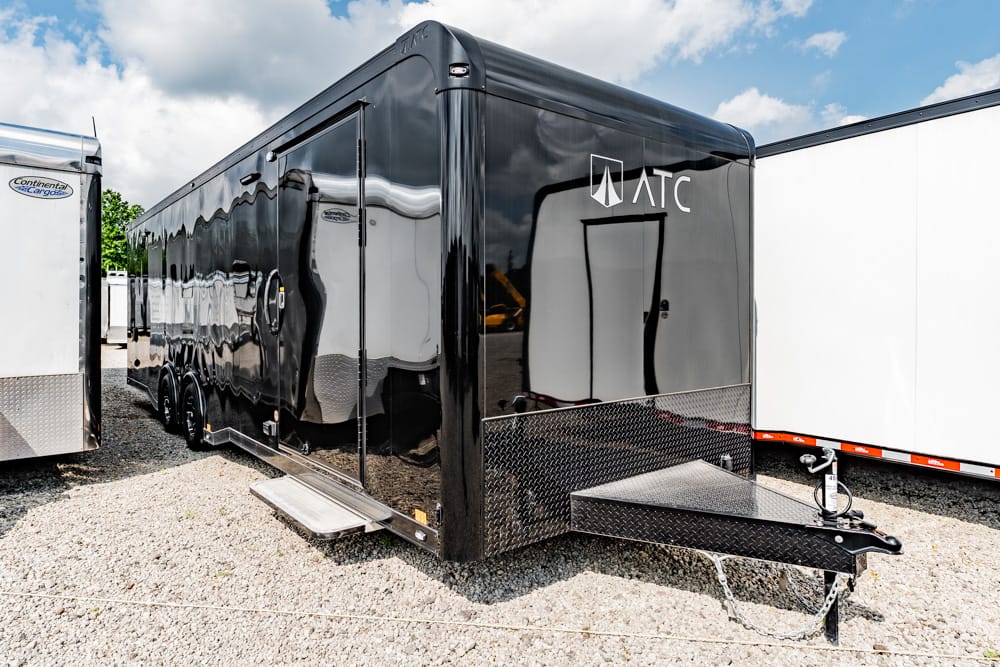 Glossy black ATC enclosed trailer with a diamond-plate front, parked on a gravel lot with a loading ramp extended.