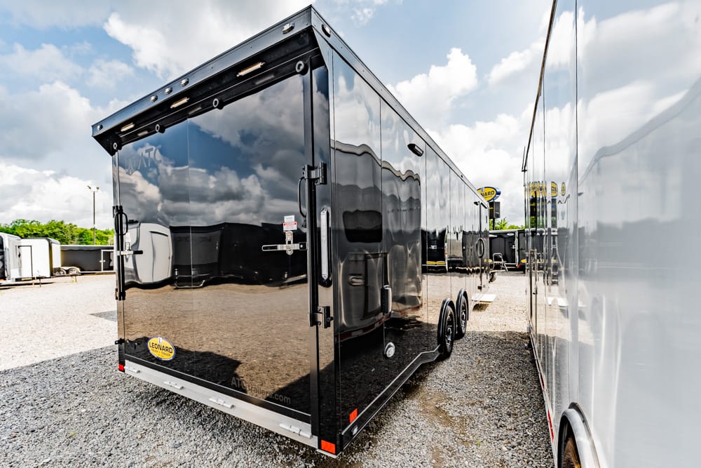 Black enclosed trailer with double rear doors on a gravel lot, reflecting nearby trailers in its glossy surface.