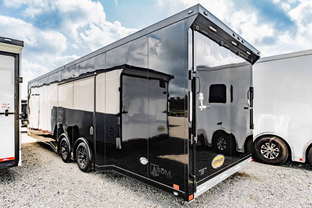 Black enclosed cargo trailer with rear ramp doors on a gravel lot, its glossy surface reflecting nearby trailers and blue sky with clouds to convey its purpose as a storage/transport unit.