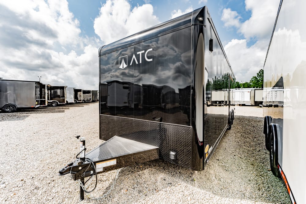 Black ATC enclosed trailer on a gravel lot, hitch at the front, diamond-plate entry area, reflected in the shiny side panel.