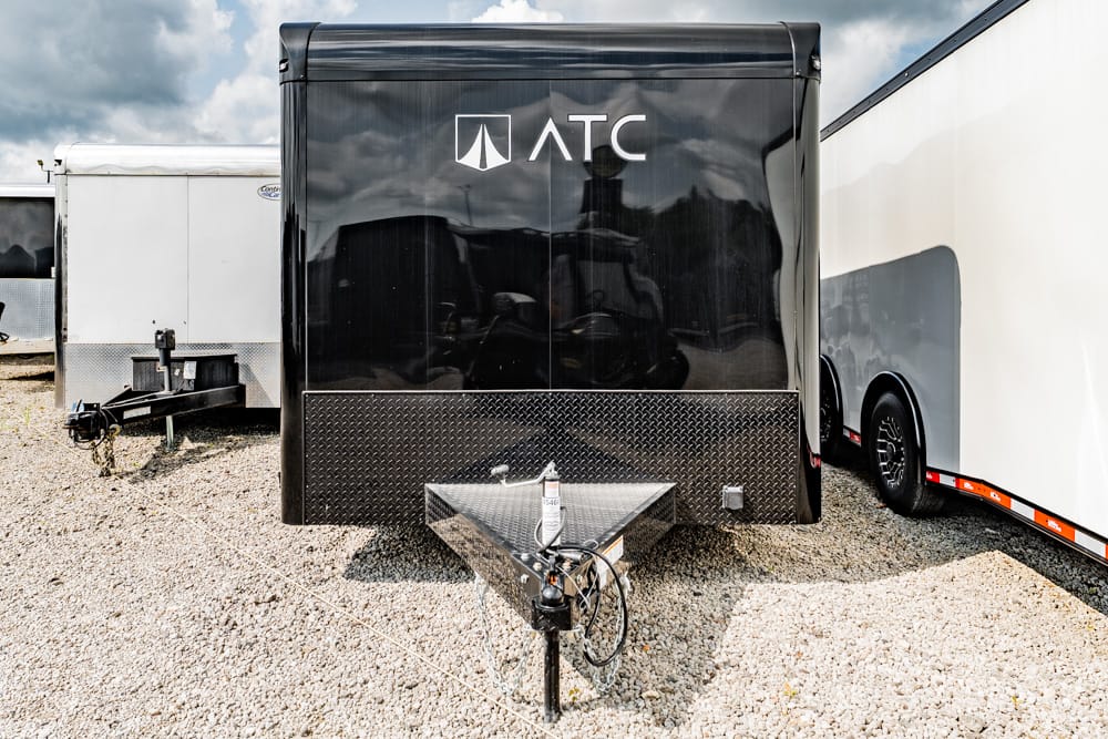 Front view of a black ATC cargo trailer with a diamond-plate lower front, hitch and jack in the gravel lot.