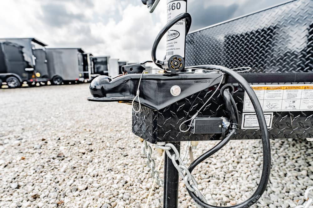 Close-up of a black trailer hitch with safety chains, wiring, and diamond-plate trailer in a gravel lot with other trailers in the background.