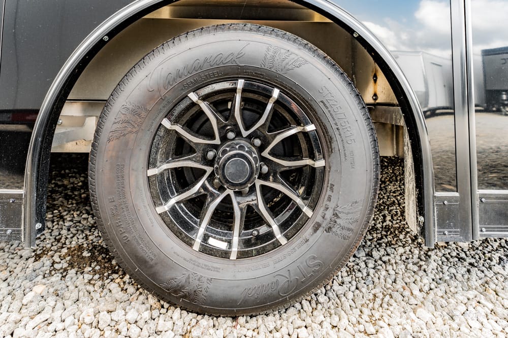 Close-up of a black alloy wheel with silver spokes mounted on a tire, resting on a gravel surface beside a dark vehicle fender.