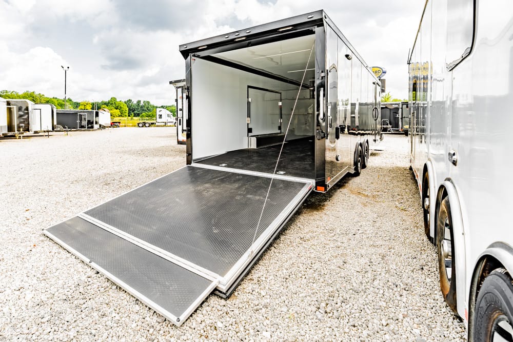 Open, empty silver cargo trailer with its loading ramp down, in a gravel lot among other trailers