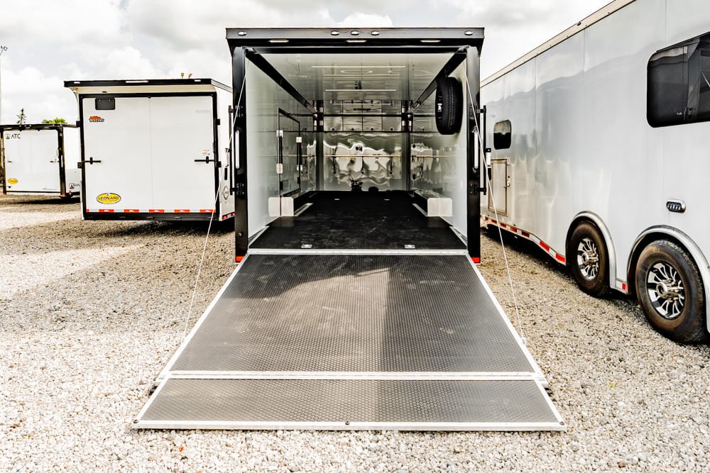 Open cargo trailer with a lowered ramp leading into an empty interior, parked among other trailers on a gravel lot.