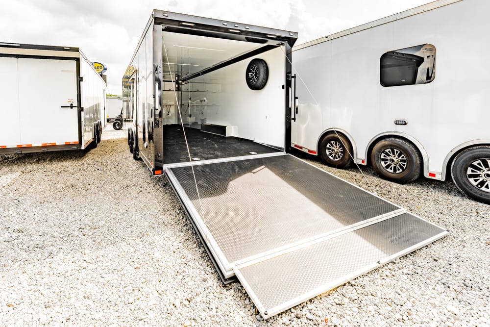 Trailer with its loading ramp lowered, ready for loading, on a gravel lot beside other enclosed trailers.