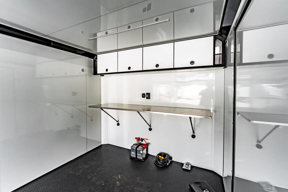 Sleek white mobile workshop interior with upper cabinets, a fold-out stainless table, and tools on the floor near hoses and batteries.
