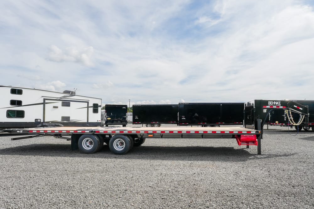 Flatbed trailer with dual axles parked on a gravel lot, red stairs at the rear and other trailers in the background.