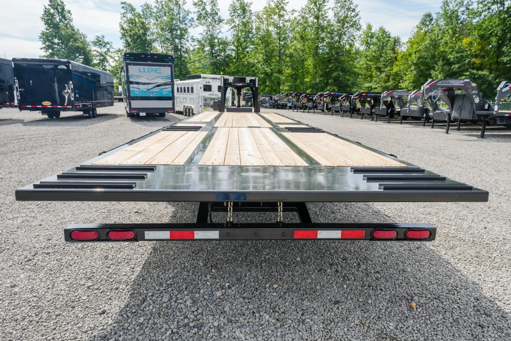 Rear view of a flatbed trailer with a wooden deck in a gravel lot, with other trailers and green trees in the background