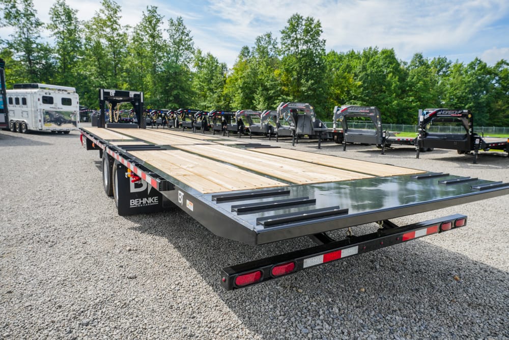 Flatbed trailer with a wooden deck and steel ramps, parked on a gravel lot with other trailers in the background, under a blue sky.