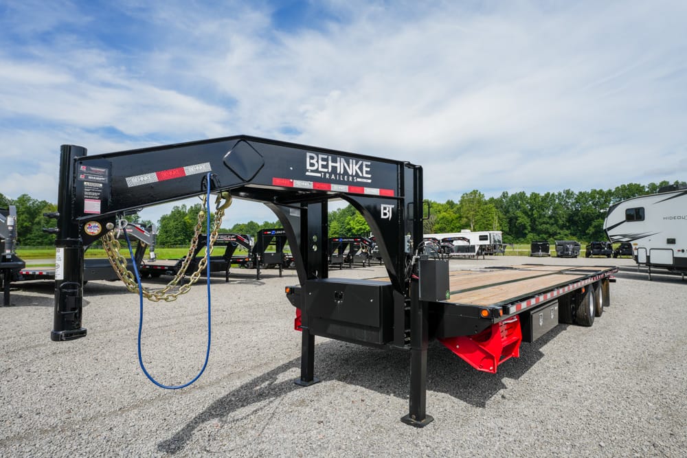 Gooseneck utility trailer in a gravel lot with a Behnke Trailers logo, safety chains, and a blue power cord hanging from the hitch; long flatbed with wooden deck and red support components visible under it.