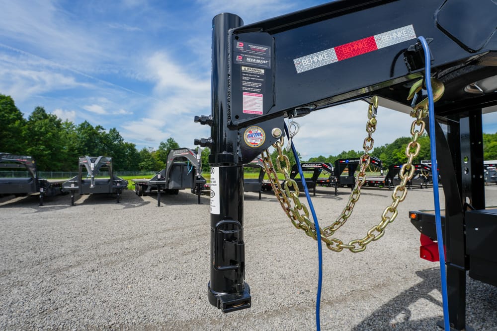 Close-up of a black trailer hitch with gold safety chains and a blue electrical cord, on a gravel lot with multiple trailers in the background under a blue sky.