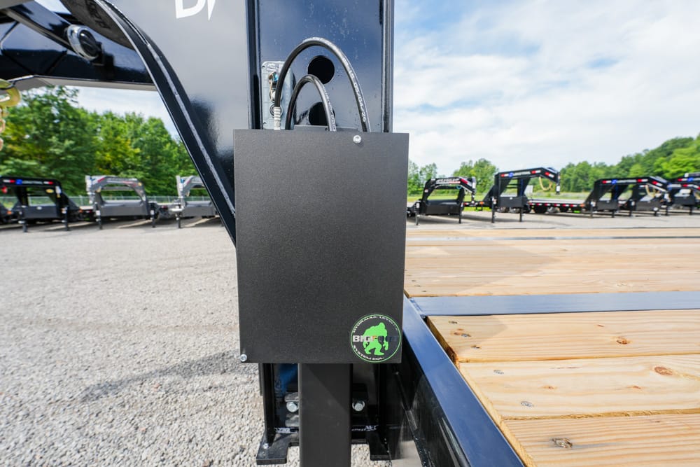 Black metal lockbox mounted on a trailer hitch of a flatbed trailer, featuring a green circular sticker, in a lot with other trailers.
