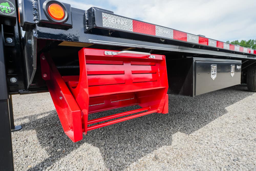 Red metal tool chest mounted under a black flatbed trailer on a gravel surface.