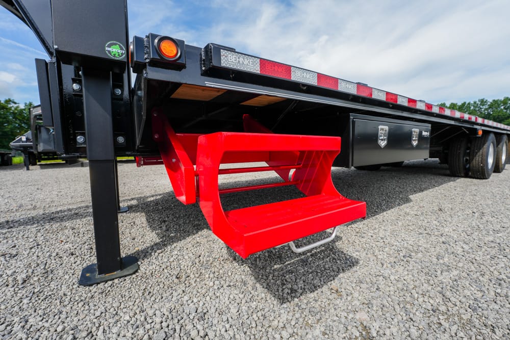 Red metal access step hanging under a black flatbed trailer on a gravel lot.