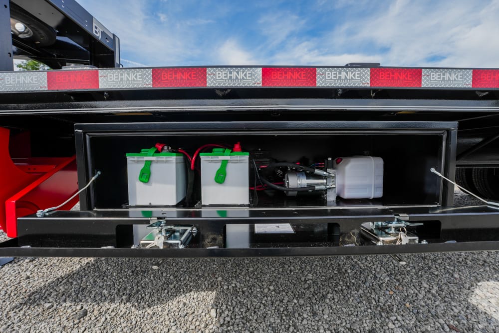 Open storage compartment on a trailer showing two white batteries with green handles and red/black wiring, plus a white reservoir, inside a black metal box.