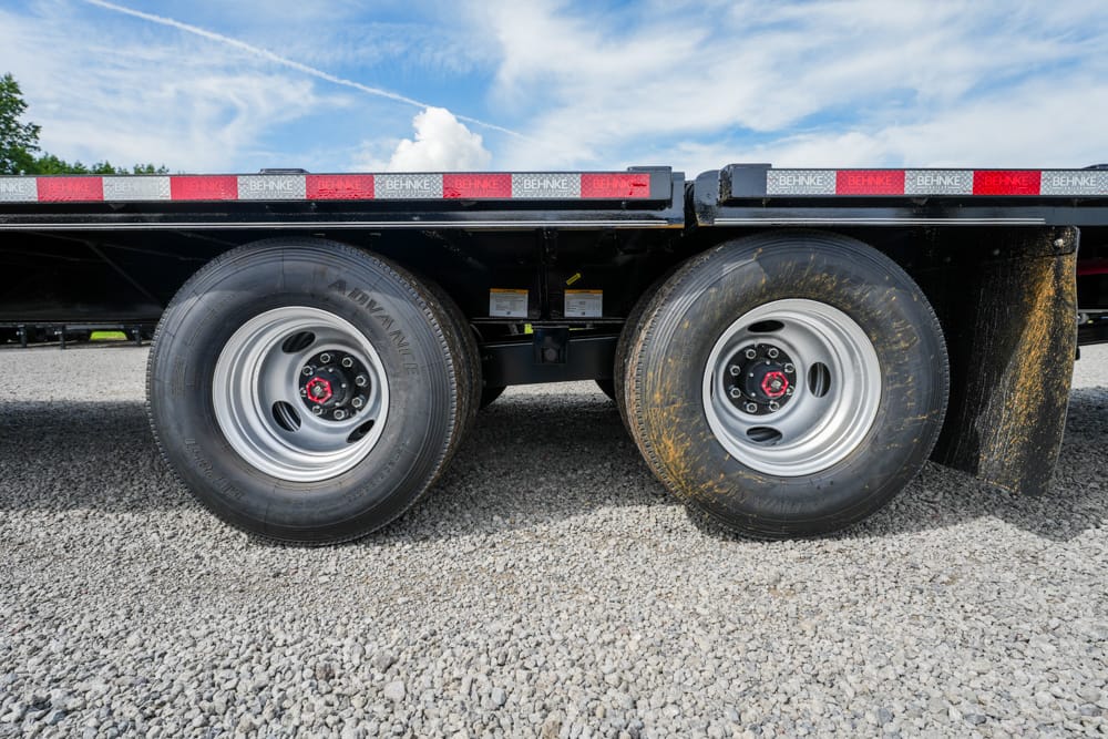 Close-up of dual rear wheels on a flatbed trailer parked on gravel under a blue sky with clouds