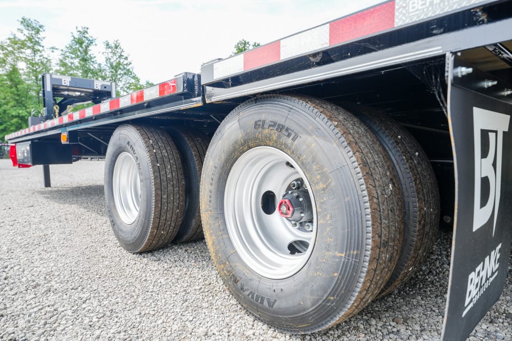Close-up of dual rear axles with tires on a heavy-duty flatbed trailer, parked on gravel.