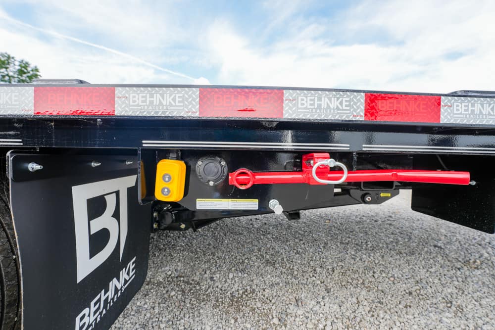 Close-up of a black trailer frame showing a red safety bar with a locking ring and a yellow control box, on a gravel lot with Behnke Trailers branding.