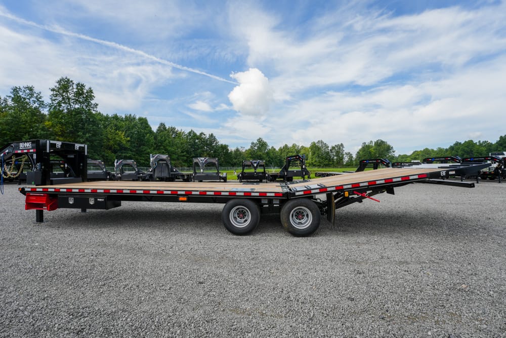 Flatbed trailer with dual rear axles on a gravel lot, with other trailers and green trees under a blue sky.