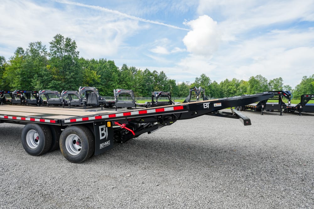 Long black flatbed trailer with dual rear axles and an extendable front section, parked on a gravel lot with green trees in the background.