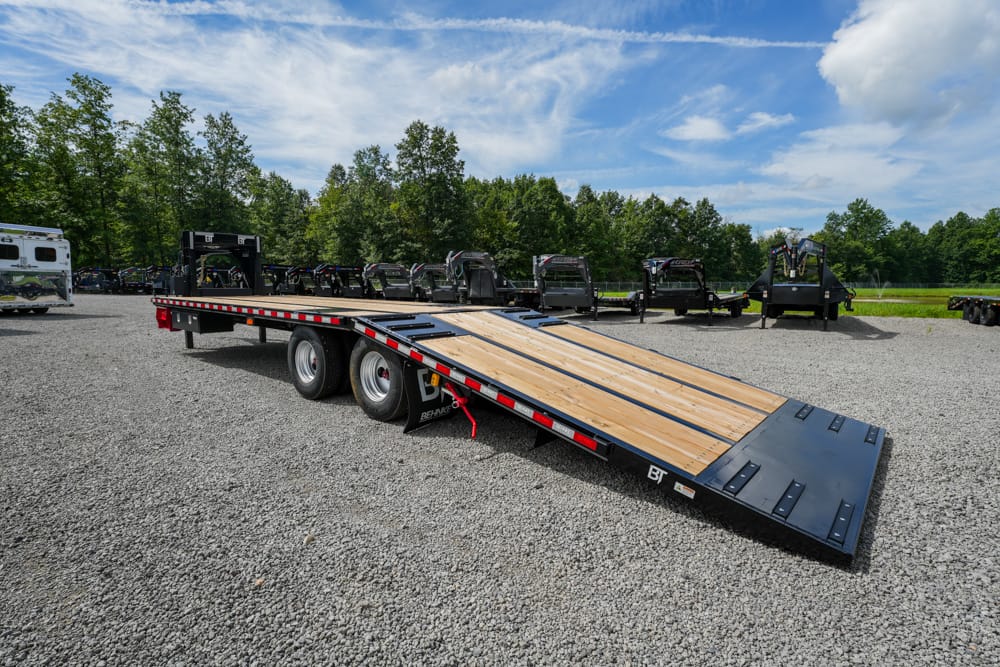 Flatbed trailer with a wooden deck parked on a gravel lot, with trees and other trailers in the background.