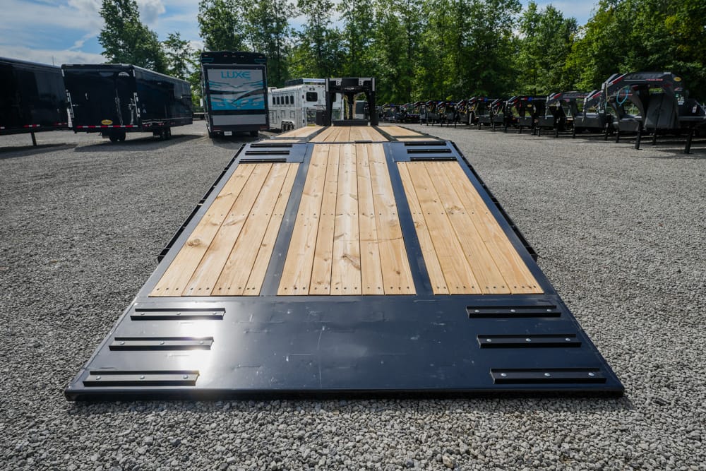 Long flatbed trailer with a wooden deck parked on a gravel lot, with other trailers and vehicles in the background