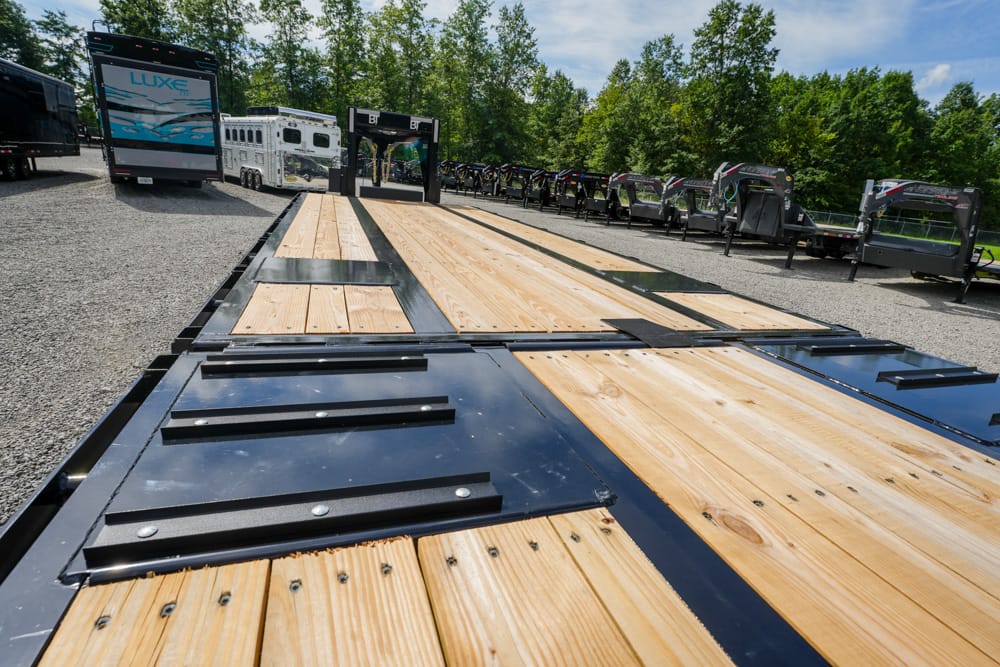 Close-up of a wooden flatbed trailer deck with black metal braces, in a gravel lot with other trailers in the background.]}}‍? Wait ensure proper JSON array.