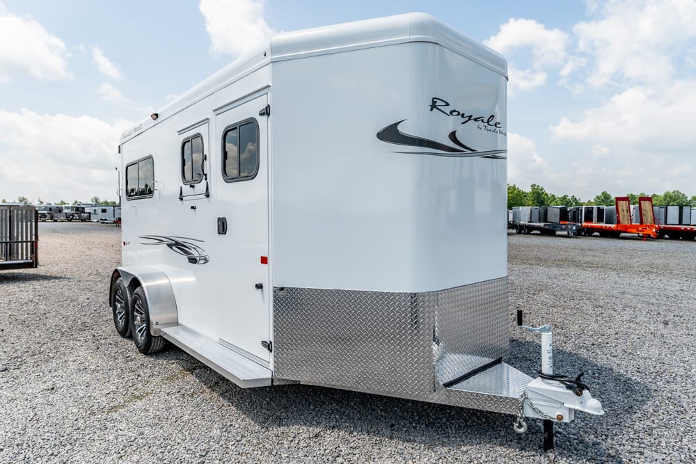White enclosed horse trailer with diamond-plate front, parked on a gravel lot; tandem axles and side windows visible.