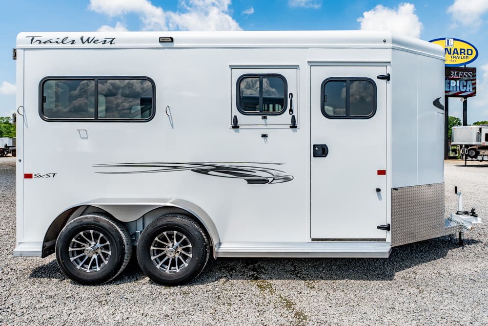 Side view of a white two-axle horse trailer with decorative gray graphics, parked on a gravel lot; visible windows and a side door with a small window and latch.