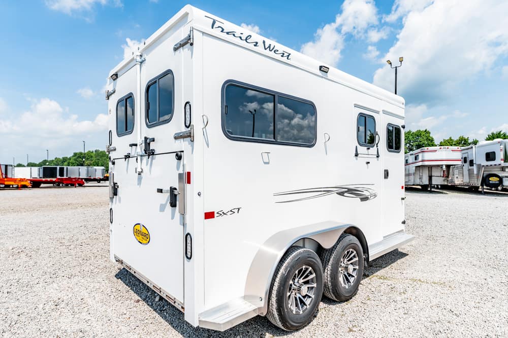 White double-axle horse trailer with Trails West branding, parked on a gravel lot under a blue sky with other trailers nearby