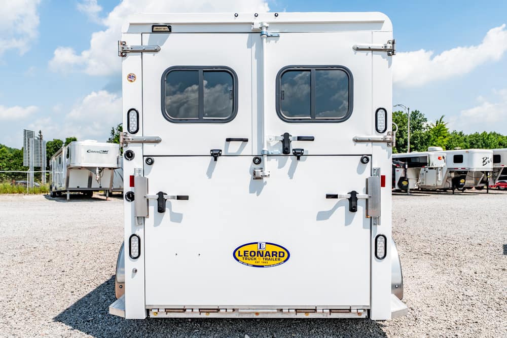 Rear view of a white Leonard trailer in a gravel lot, showing two windows, metal latches, and a Leonard logo on the bumper under a blue sky.