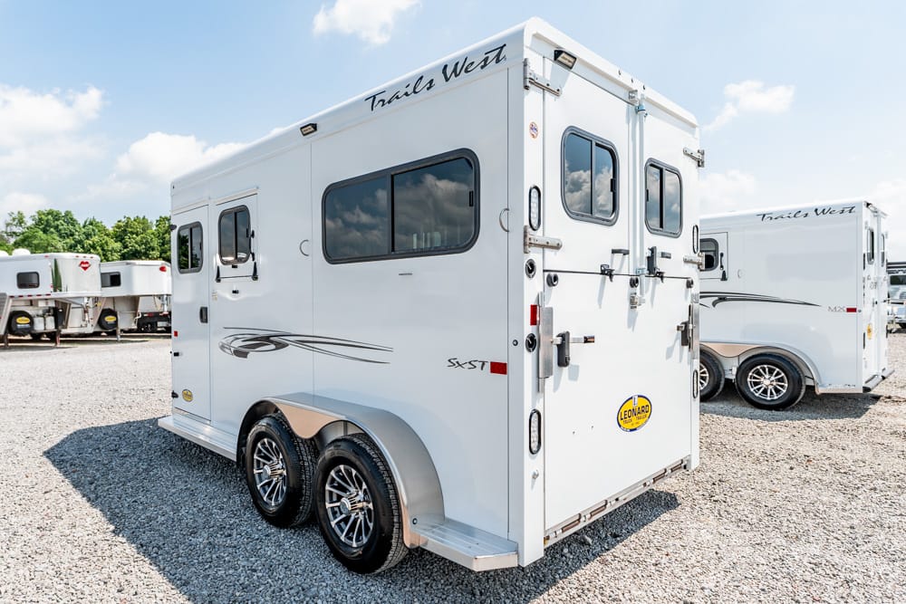 White Trails West horse trailer with tandem wheels and rear double doors, parked on a gravel lot with other trailers in the background.