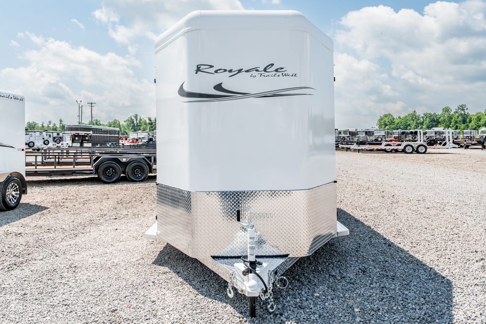 White enclosed horse trailer with diamond-plate front, hitch and chains, parked on a gravel lot with other trailers nearby under a blue sky