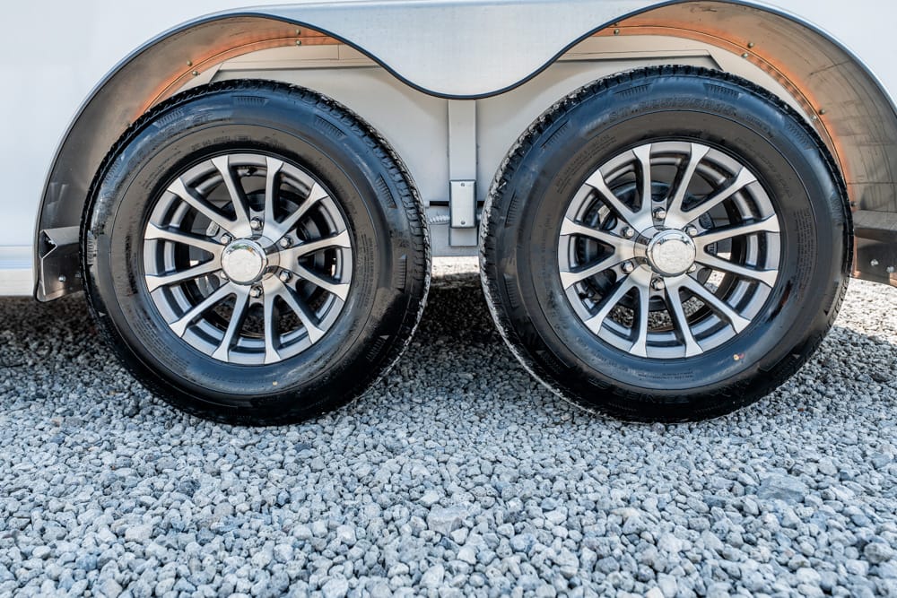 Dual trailer wheels with silver alloy rims on a gravel surface, showing tire treads and wheel hubs above the ground clearance.