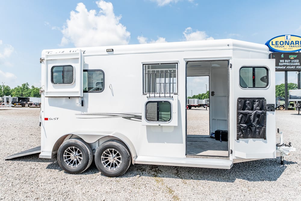 White horse trailer on a gravel lot with two axles, side entry door open and rear ramp lowered for loading horses or gear.