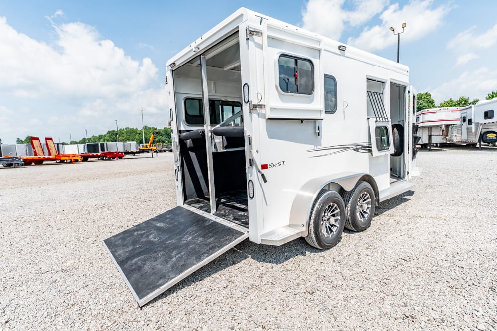 White horse trailer with ramp down, showing interior stalls on a gravel lot already loaded for transport.