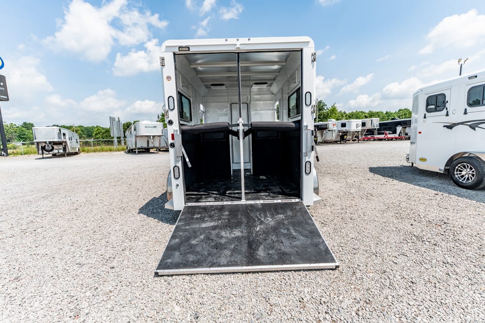 Open white horse trailer with ramp lowered, showing two divider stalls and seating inside on a gravel lot.