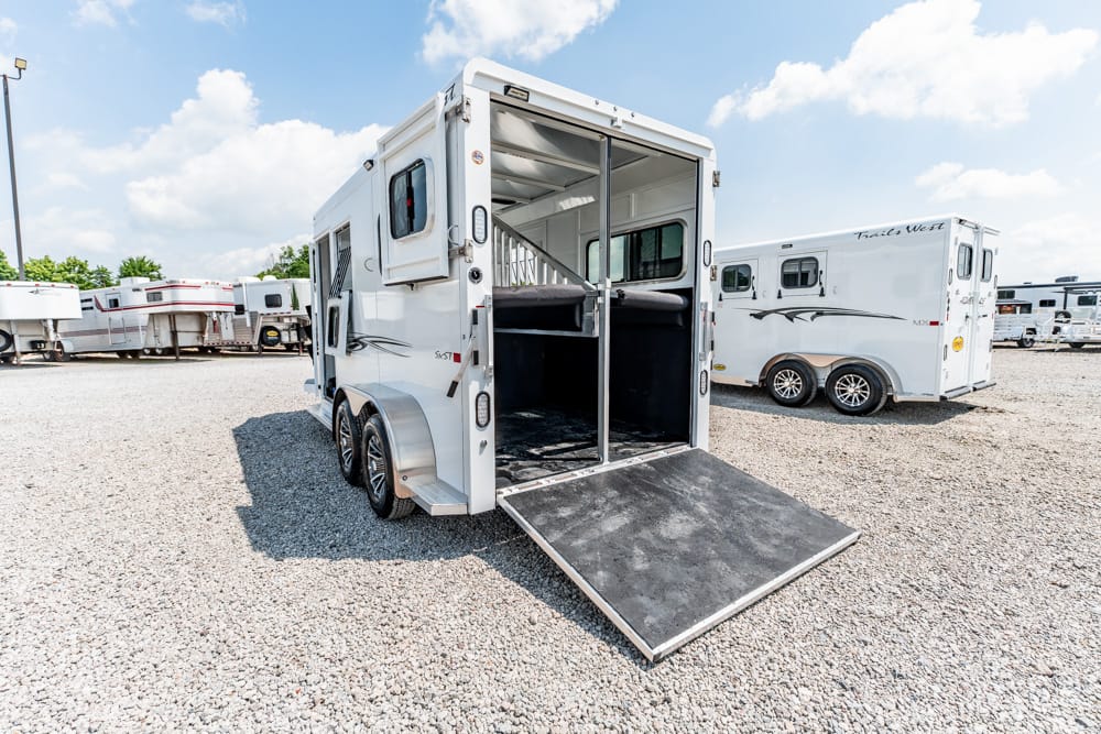 White two-axle horse trailer with the ramp lowered, parked on a gravel lot with other trailers nearby
