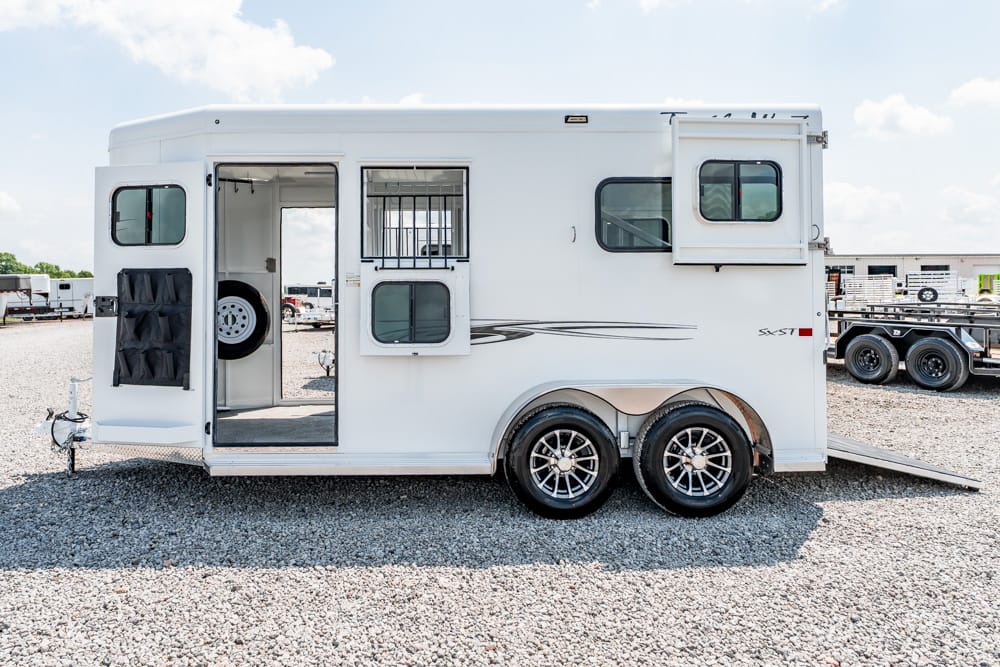 White horse trailer with two axles, side door open and spare tire inside on a gravel lot