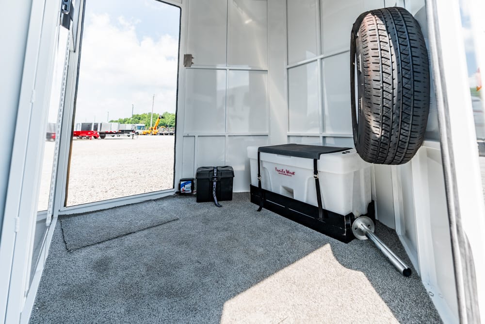Interior of a trailer with a mounted spare tire, a white cooler on a rack, and tools near an open door to a gravel lot.