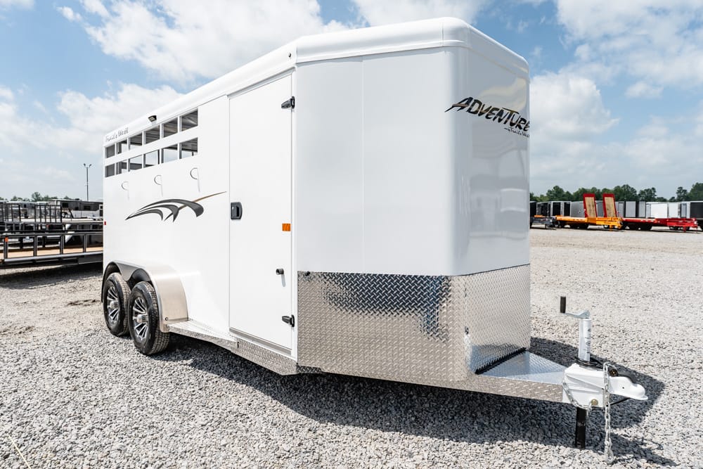 White two-axle horse trailer with diamond-plate front, parked on gravel, hitch at the prow and 'Adventure' branding on the side/upper front.