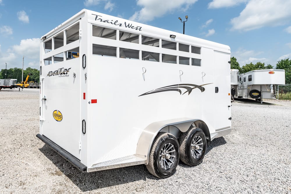 Rear and side view of a white Trails West horse trailer with dual axles and decorative swoosh graphics on a gravel lot.
