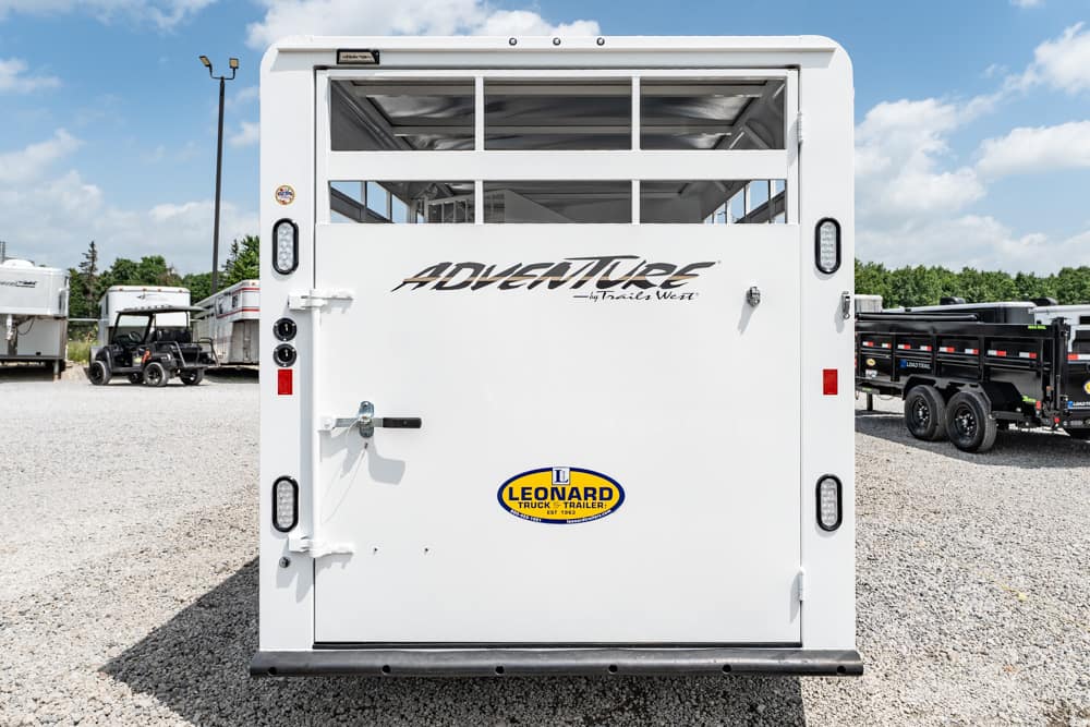 Back of a white trailer with 'ADVENTURE by Trails West' branding in a dealership lot, Leonard trailer sticker visible on the door.